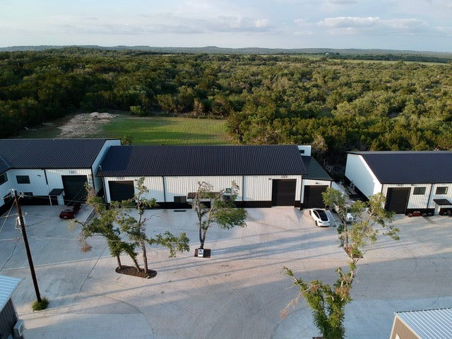 The image shows an aerial view of a group of white industrial buildings with black roofs and black trim on the doors. The buildings is the AJ Networx US and has a paved parking lot in front. The buildings are surrounded by a vast expanse of trees in the background. A few cars are parked in the lot. The image is shot on a bright, sunny day. There is a small tree to the left of the image, and another tree sits in the foreground, The top of a small red car, the partial view of a white car.