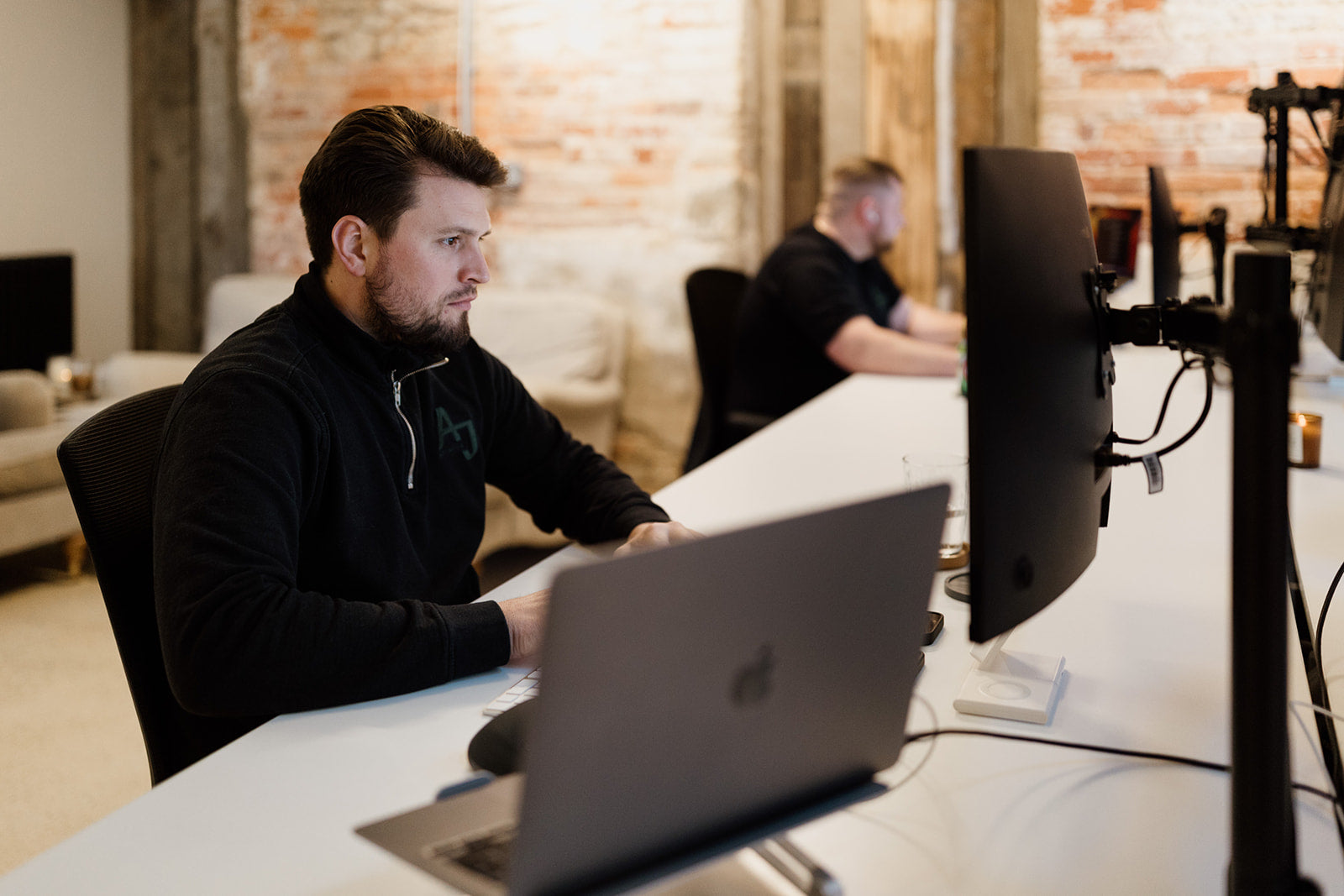  The image shows a focused man working at his laptop in a modern office with exposed brick walls. Another man works at a desk behind him. Both wear dark sweaters. The space is equipped with large monitors and tech accessories, blending rustic charm with a contemporary, productive atmosphere.