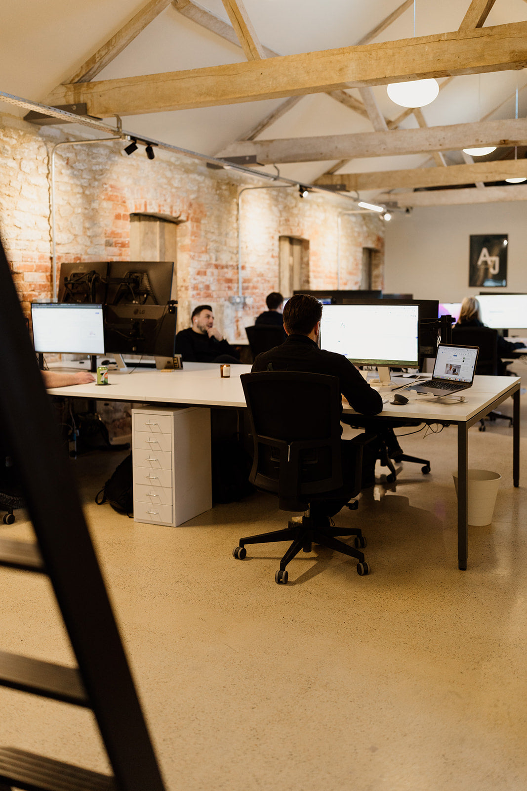 The room features exposed brick walls and wooden beams on the ceiling. Several people are working at large, white desks equipped with multiple monitors and laptops. 