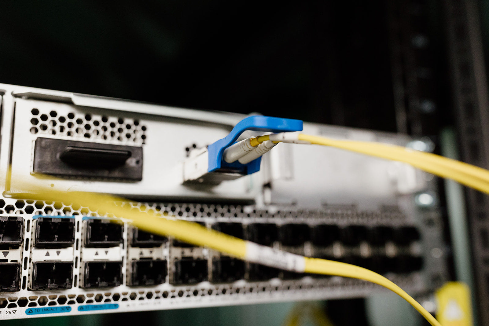  A image of a close-up of a server rack with yellow fiber optic cables plugged into ports on the back. The cables are connected to a blue and white connector.