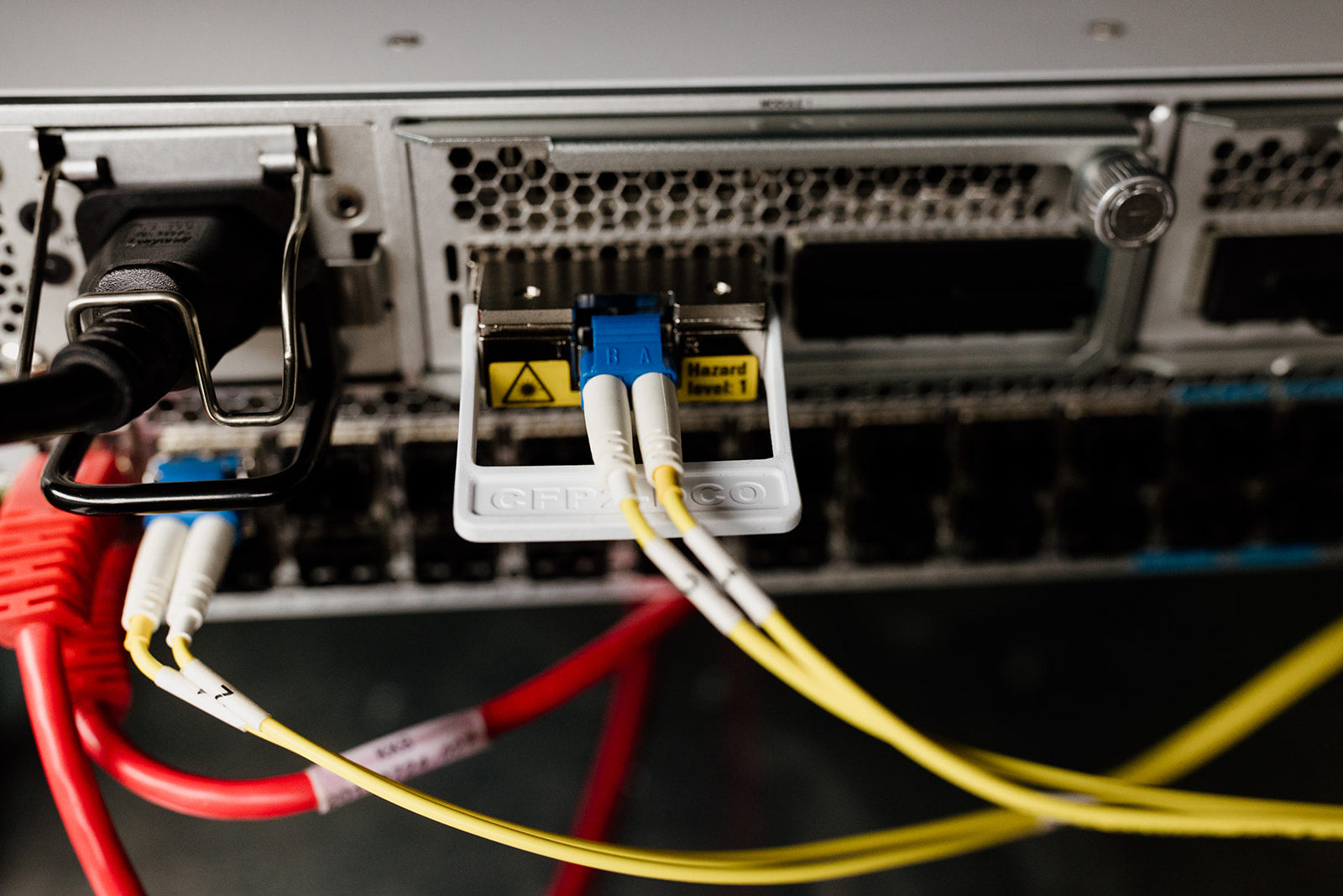 A close-up of a server rack with multiple network cables plugged in. The focus is on two yellow fiber optic cables that are connected to a blue transceiver. The cables are running across the rack, which is filled with a series of network ports. The background is blurred, which further emphasizes the cables and transceiver. 