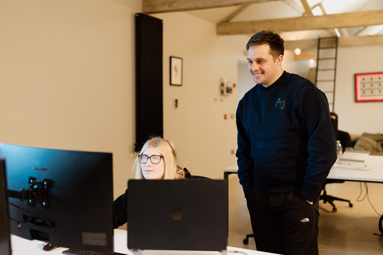 The image shows a casual office setting with two people interacting. A woman with blonde hair and glasses is seated at a desk, focused on her computer monitor. A man, standing beside her, is smiling and appears to be engaged in conversation. Both are wearing dark sweaters, with the man’s sweater displaying an "AJ" logo. 